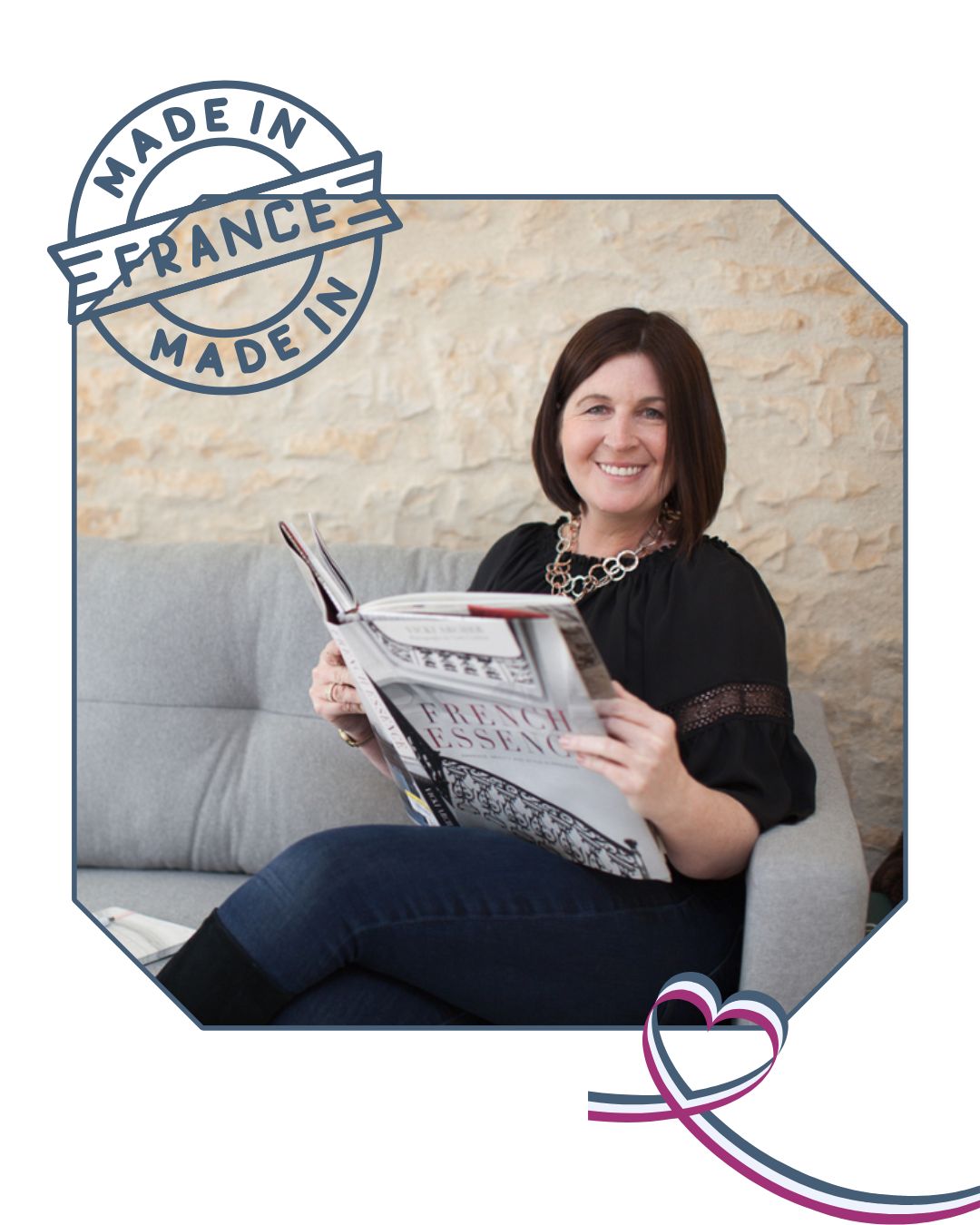 A smiling woman sits on a light gray couch, holding an open book titled 'French Essence' with decorative black and white designs on the cover. She wears a black blouse with lace details and a chunky silver necklace. The image includes a 'Made in France' stamp in the top left and a heart-shaped ribbon design in the bottom right corner.