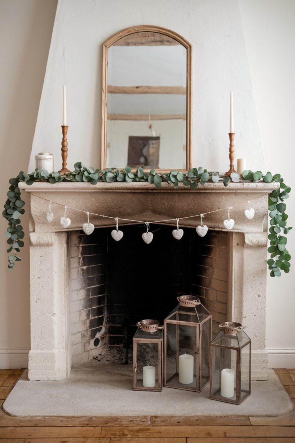 Simple yet chic Valentine’s fireplace decor featuring white ceramic hearts strung across a eucalyptus garland with wooden lanterns and candles.